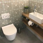 Stock Photo of modern white luxury washroom / bathroom cloakroom WC suite with contemporary curved wall hung toilet pan hanging on washroom wall, small rectangular sink basin, single chrome mixer tap on wooden shelf surface, toilet roll, brush, patterned tiles